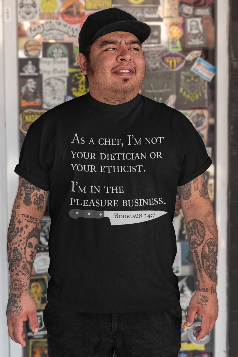 May include: Black t-shirt with white text that reads "As a chef, I'm not your dietician or your ethicist. I'm in the pleasure business. Bourdain 24/7". A white chef's knife is pictured below the text.