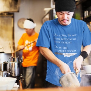 May include: A chef wearing a blue t-shirt with the text "As a chef, I'm not your dietician or your ethicist. I'm in the pleasure business. Boudin 247" is preparing food in a kitchen.