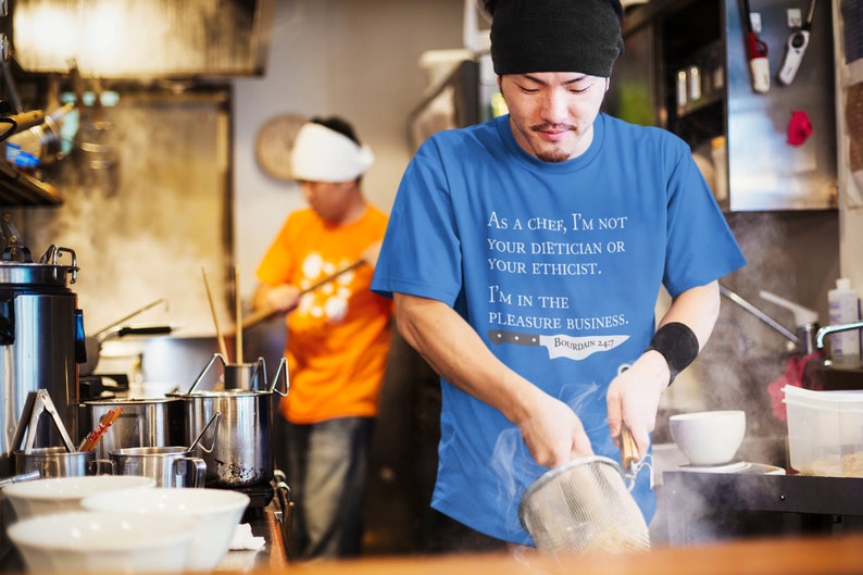 May include: A chef wearing a blue t-shirt with the text "As a chef, I'm not your dietician or your ethicist. I'm in the pleasure business. Boudin 247" is preparing food in a kitchen.