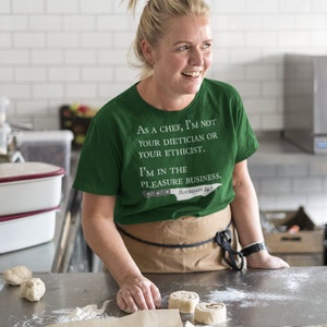 May include: A woman wearing a green t-shirt with the text "As a chef, I'm not your dietician or your ethicist. I'm in the pleasure business. Bourdain 24/7" is preparing cinnamon rolls in a kitchen.