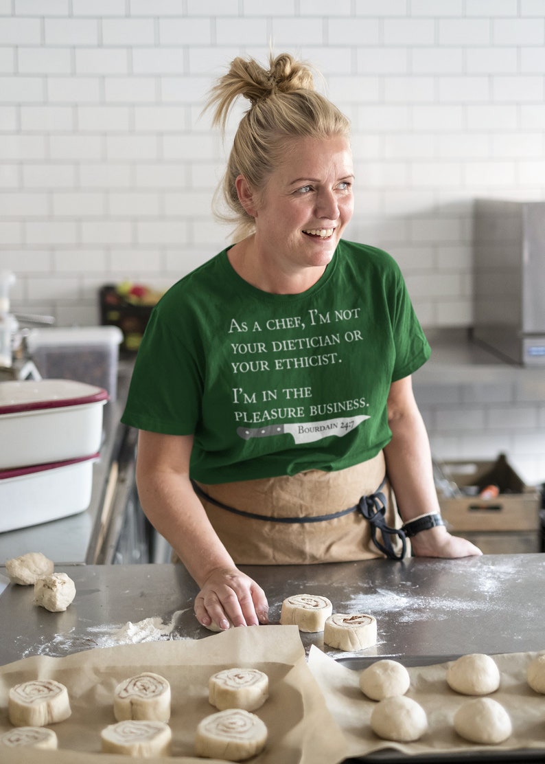 May include: A woman wearing a green t-shirt with the text "As a chef, I'm not your dietician or your ethicist. I'm in the pleasure business. Bourdain 24/7" is preparing cinnamon rolls in a kitchen.