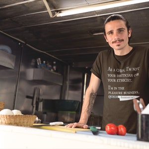 May include: A brown t-shirt with white text that reads "As a chef, I'm not your dietician or your ethicist. I'm in the pleasure business."  The text is accompanied by a small knife graphic with the text "Bourdain 147".