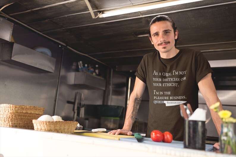 May include: A brown t-shirt with white text that reads "As a chef, I'm not your dietician or your ethicist. I'm in the pleasure business."  The text is accompanied by a small knife graphic with the text "Bourdain 147".