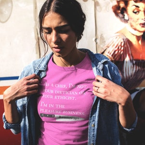 May include: A pink t-shirt with a white graphic print that reads "As a chef, I'm not your dietician or your ethicist. I'm in the pleasure business."  The graphic also includes a knife with the text "Bowman 4:17".