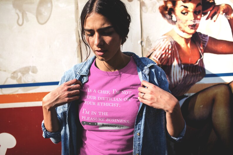 May include: A pink t-shirt with a white graphic print that reads "As a chef, I'm not your dietician or your ethicist. I'm in the pleasure business."  The graphic also includes a knife with the text "Bowman 4:17".