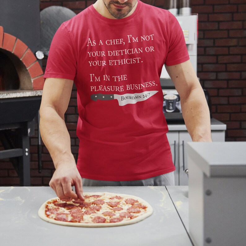 May include: A red t-shirt with the text "As a chef, I'm not your dietician or your ethicist. I'm in the pleasure business. Bourdain 24/7" and a knife graphic. A person is placing pepperoni on a pizza.