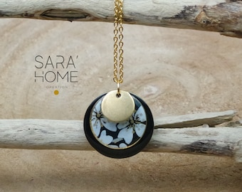 May include: A gold-colored chain necklace with a pendant featuring a black and white floral design. The pendant has a gold-colored disc on top and is set against a natural wood background. The text "SARA' HOME CREATION" is visible.