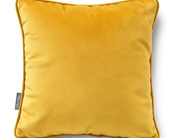 May include: A square mustard yellow velvet throw pillow with piped edges. The pillow features a small grey fabric tag on the left side with the text Celina Digby printed in white. The surface has a soft, uniform texture throughout.