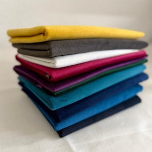 May include: A pile of folded fabric swatches in a range of colours: yellow, grey, white, magenta, purple, teal, and navy. The fabrics look soft and velvety, possibly velvet or suede.