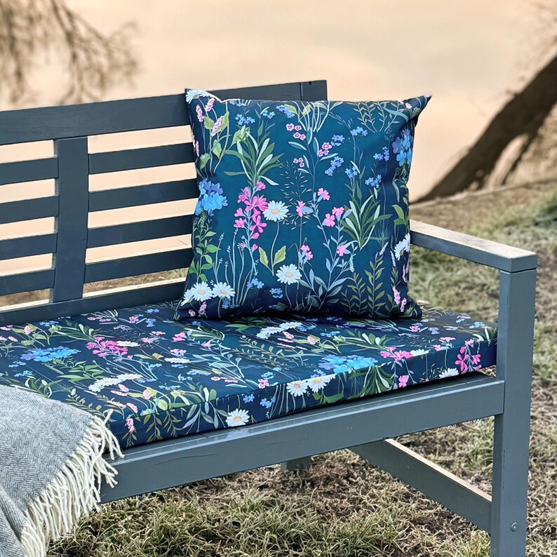 May include: A blue outdoor bench with a floral patterned cushion and a matching throw pillow. The cushion and pillow feature a dark blue background with pink, white, and blue flowers.