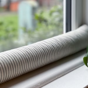 May include: A white and black striped fabric draught excluder for a window or door. The draught excluder is rolled up and placed on a white window sill.