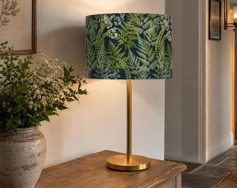 May include: A table lamp with a cylindrical lampshade featuring a dark green and light green fern leaf pattern. The lamp has a slender, polished gold-tone metal stem and a matching circular base, emitting a warm glow from beneath the shade.