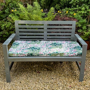 May include: A grey wooden bench with a floral patterned cushion. The bench is sitting on a gravel surface.