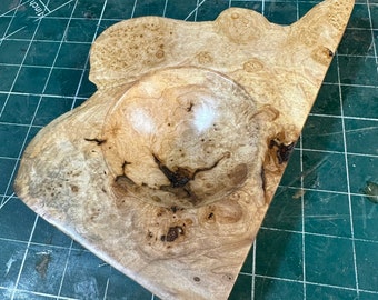 Könnte beinhalten: Eine Holzschaale mit einer natürlichen, unregelmäßigen Form und einem tiefen, runden Innenraum. Die Schale hat eine hellbraune Farbe mit dunkelbraunen Streifen und Knoten. Die Schale steht auf einer grünen Schneidematte.