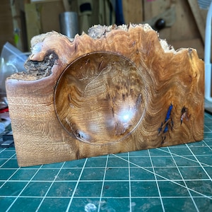 Puede incluir: Un cuenco de madera con un borde natural y un interior liso y redondeado. El cuenco está hecho de madera marrón claro con vetas marrón oscuro y una incrustación de resina azul.