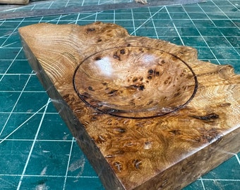 Könnte beinhalten: Eine Holzschale mit einer kreisförmigen Vertiefung, hergestellt aus einem Wurzelholz mit einer satten braunen und goldenen Farbe. Die Schale liegt auf einer grünen Schneidematte.