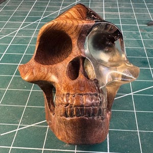 May include: A handcrafted wooden skull sculpture with a clear, resin-filled eye socket. The skull is carved from dark brown wood, showcasing the natural grain. The sculpture is on a green cutting mat.