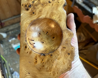 Könnte beinhalten: Eine handgefertigte Holzschale mit natürlicher, hellbrauner Oberfläche. Die Schale hat eine kreisförmige Vertiefung und eine einzigartige, organische Form. Die Holzmaserung ist sichtbar und trägt zum rustikalen Charme dieses Dekorationsstücks bei.