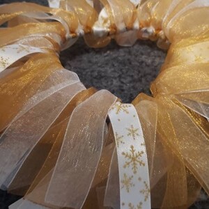 May include: A gold and white wreath made of multiple layers of sheer ribbon. The ribbon has a snowflake pattern.