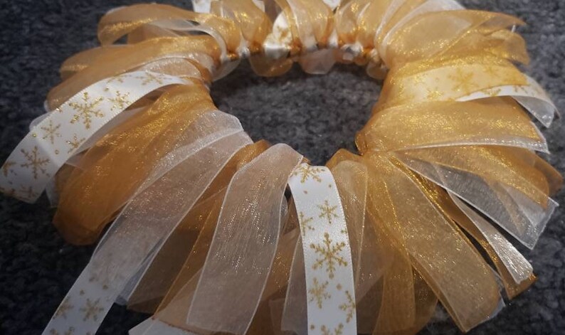 May include: A gold and white wreath made of multiple layers of sheer ribbon. The ribbon has a snowflake pattern.