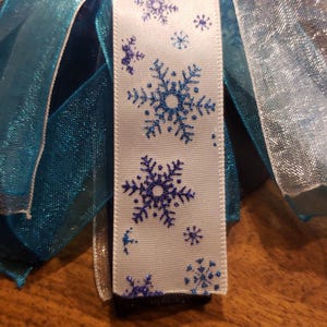 May include: White ribbon with blue glitter snowflakes printed on it. The ribbon is surrounded by blue and white sheer ribbon.