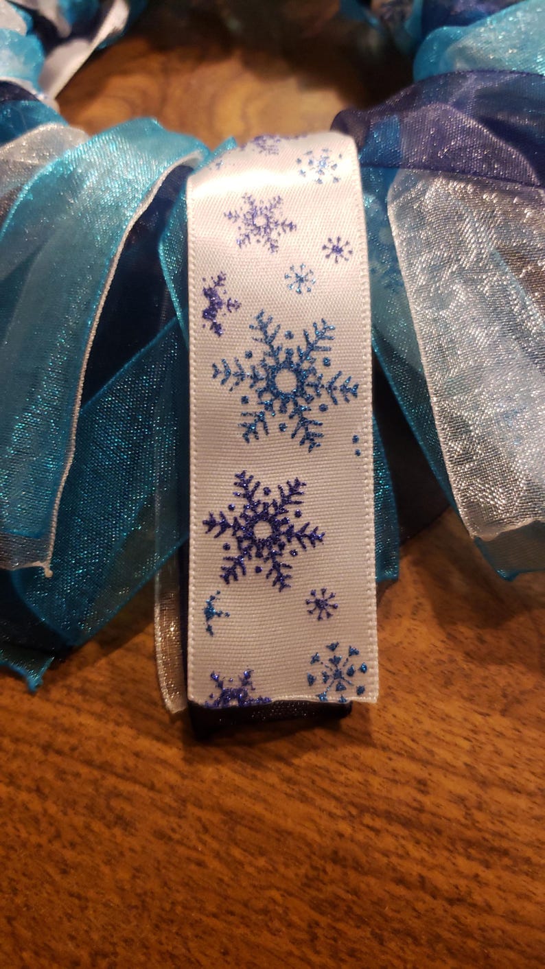 May include: White ribbon with blue glitter snowflakes printed on it. The ribbon is surrounded by blue and white sheer ribbon.