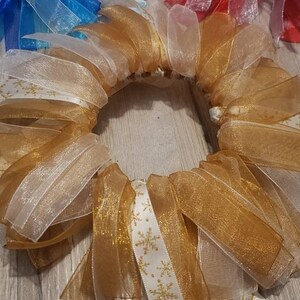 May include: A gold and white wreath made of sheer ribbon with a snowflake pattern. The wreath is made of multiple layers of ribbon and is tied together in the back.