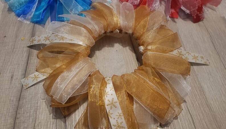 May include: A gold and white wreath made of sheer ribbon with a snowflake pattern. The wreath is made of multiple layers of ribbon and is tied together in the back.