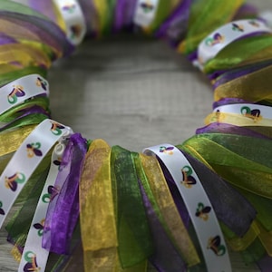 May include: A Mardi Gras wreath made with purple, green, and gold ribbon. The ribbon is decorated with white fleur-de-lis.
