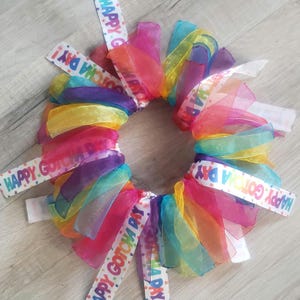 May include: A rainbow-colored tulle wreath with white ribbon streamers that say "Happy Gotcha Day!"