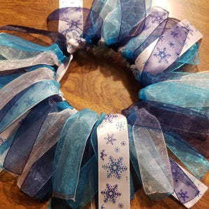 May include: A blue, white, and silver snowflake wreath made of ribbon. The ribbon is layered and ruffled to create a full and festive look.