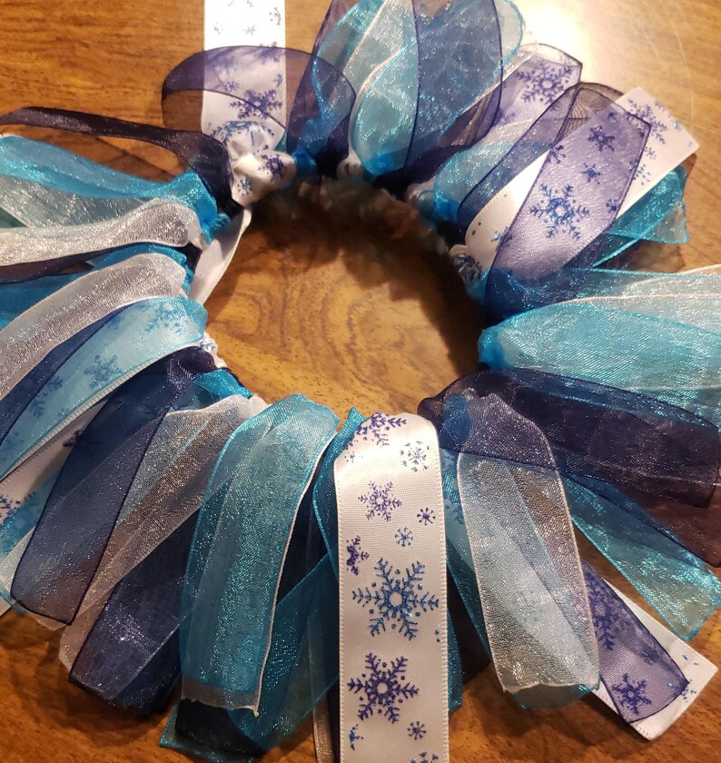 May include: A blue, white, and silver snowflake wreath made of ribbon. The ribbon is layered and ruffled to create a full and festive look.