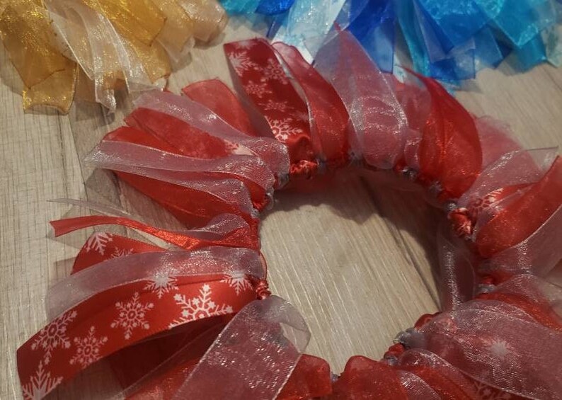 May include: A red and white ribbon wreath with snowflake patterns. The wreath is made of multiple layers of sheer ribbon and is tied together with a white ribbon.