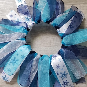 May include: A blue, white, and silver tulle wreath with snowflake patterns. The wreath is made of multiple layers of tulle ribbon.