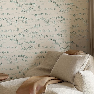 May include: A cozy living room scene featuring a cream-colored, textured armchair with a tan throw blanket and pillows. The backdrop is a wallpaper with a green mountain and tree pattern. A small wooden side table holds books.