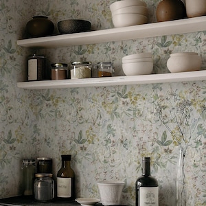 Puede incluir: Una escena de cocina con estantes blancos que exhiben varios cuencos de cerámica, frascos y botellas. Los estantes están contra un papel pintado floral. Una botella de aceite de oliva y un jarrón con flores secas están en el mostrador.