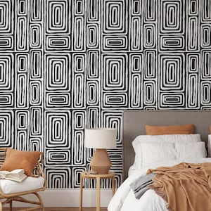 Puede incluir: Papel pintado geométrico en blanco y negro con un patrón repetitivo de formas rectangulares. El papel pintado es el telón de fondo de una escena de dormitorio con una cama, una silla y una mesita de noche. La cama tiene ropa de cama blanca y una manta marrón.