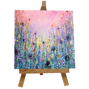 May include: A small canvas painting on an easel depicting a field of colourful wildflowers with a pink and purple sky in the background.