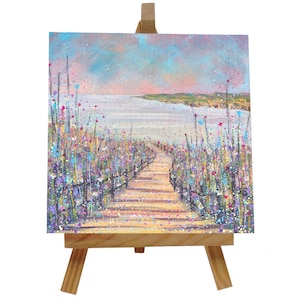 Könnte beinhalten: Original Acrylgemälde auf Leinwand, das eine Küstenlandschaft zeigt. Ein Holzsteg führt zum Meer, flankiert von bunten Wildblumen. Der Himmel wechselt von Rosa zu Blau und schafft eine ruhige Landschaft. Das Gemälde wird auf einer kleinen Holzstaffelei präsentiert.