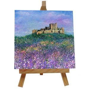 May include: A square acrylic painting depicting a castle on a green hill, with a field of purple and pink flowers in the foreground. The sky is a blend of blue and purple. The artwork is presented on a small wooden easel.