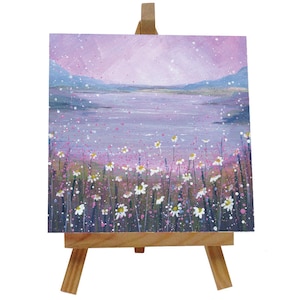 Puede incluir: Pequeña pintura cuadrada sobre caballete de madera. La obra representa un paisaje sereno con un lago lavanda, montañas azules y un campo de margaritas blancas. El cielo es rosa pálido con motas blancas.