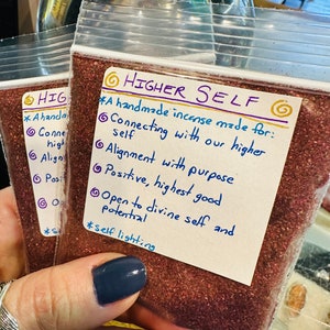 May include: A small clear plastic bag filled with red incense. The bag has a white label with the text "HIGHER SELF" and a list of benefits: "A handmade incense made for: Connecting with our higher self Alignment with purpose Positive, highest good Open to divine self and potential *self lighting"