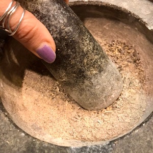 Pode incluir: Um close-up de um almofariz e pil&atilde;o de pedra cinza sendo usados para moer ervas e especiarias. O almofariz est&aacute; cheio de um p&oacute; marrom claro e pequenos peda&ccedil;os de ervas secas.