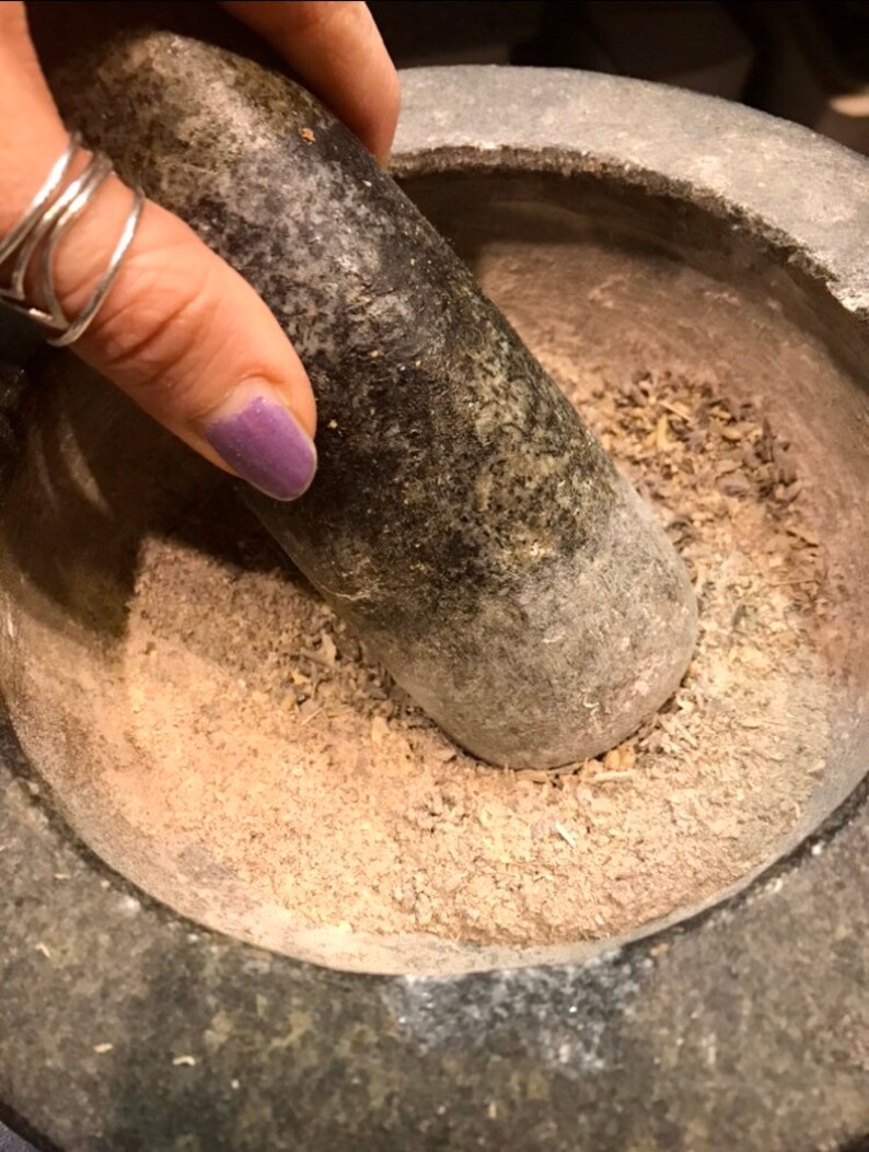 Pode incluir: Um close-up de um almofariz e pil&atilde;o de pedra cinza sendo usados para moer ervas e especiarias. O almofariz est&aacute; cheio de um p&oacute; marrom claro e pequenos peda&ccedil;os de ervas secas.