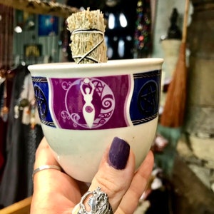 May include: A white ceramic cup with a purple, blue, and gold design featuring a woman with raised arms. The cup is holding a bundle of dried white sage smudge sticks.