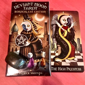 May include: A box of Deviant Moon Tarot cards, borderless edition, with a black and white illustration of a character with a crescent moon on their face. The box is on a pink surface next to a card from the deck, The High Priestess, which features a black and white illustration of a character with a crescent moon on their face. The card is on a black and white checkered surface.