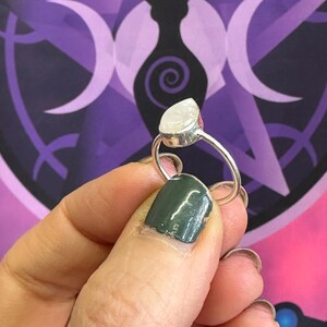 May include: A silver ring with a teardrop-shaped white gemstone. The ring is on a finger with dark green nail polish.