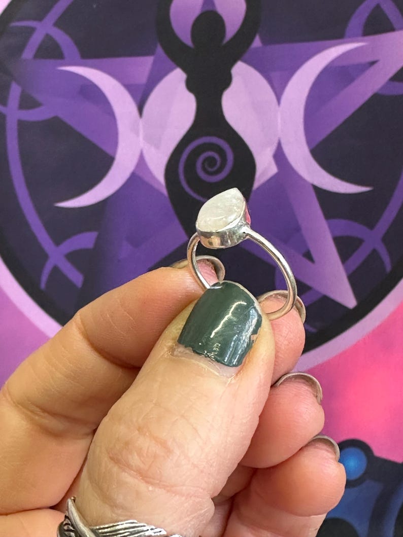 May include: A silver ring with a teardrop-shaped white gemstone. The ring is on a finger with dark green nail polish.