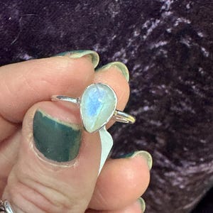 May include: A silver ring with a teardrop-shaped moonstone. The stone is iridescent and has a milky white color.