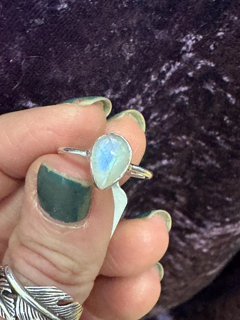May include: A silver ring with a teardrop-shaped moonstone. The stone is iridescent and has a milky white color.